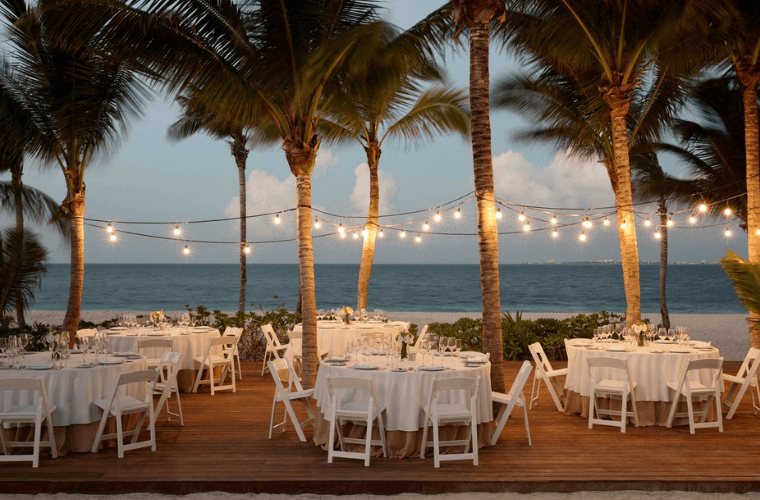 white tables and chairs in a beach wedding set up decorated with lights for illumination as the sun goes down 