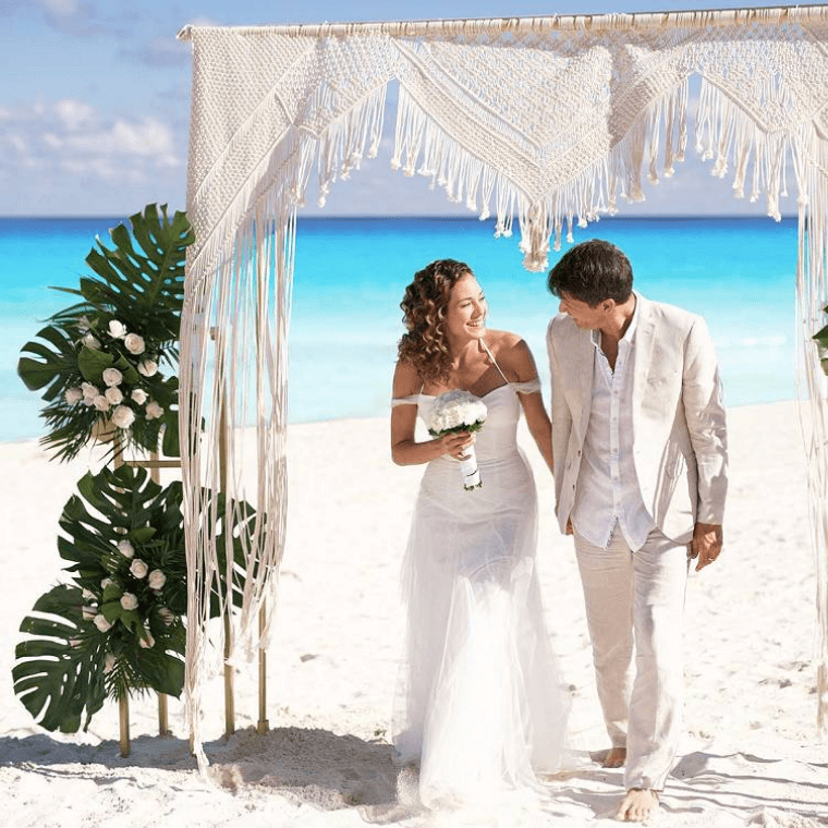 a bride and groom dressed in white standing under a wedding arch on the beach with the Caribbean Sea in the background 