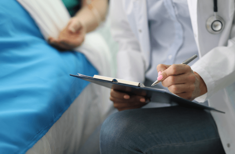 the arm of a man in a hospital bed with the hands of a doctor writing notes 