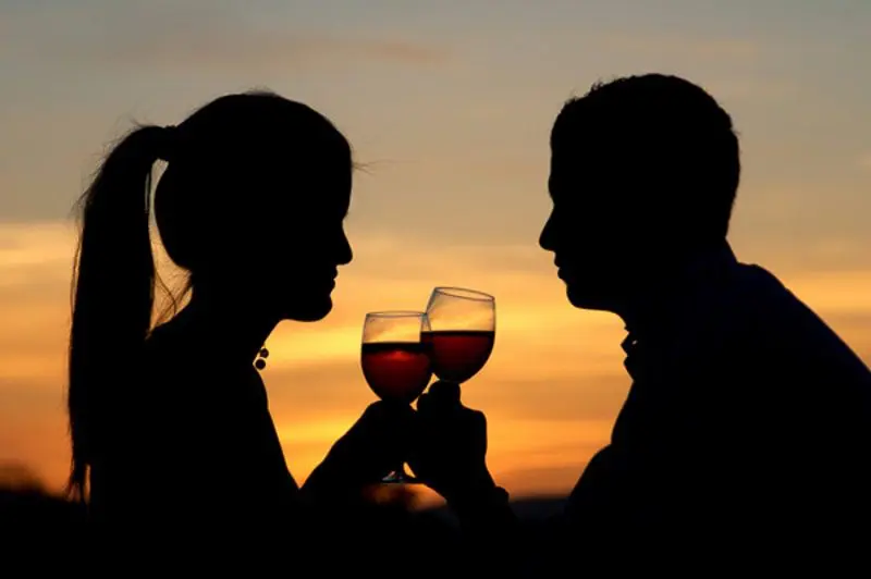 A couple romantically toasts with glasses of wine as the sun fades in the background