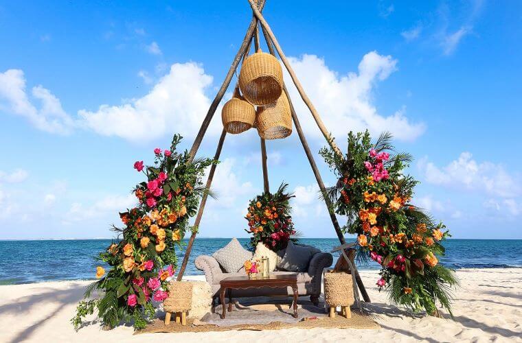 A wooden frame with floral decor and a seat below set up for a beach wedding ceremony 